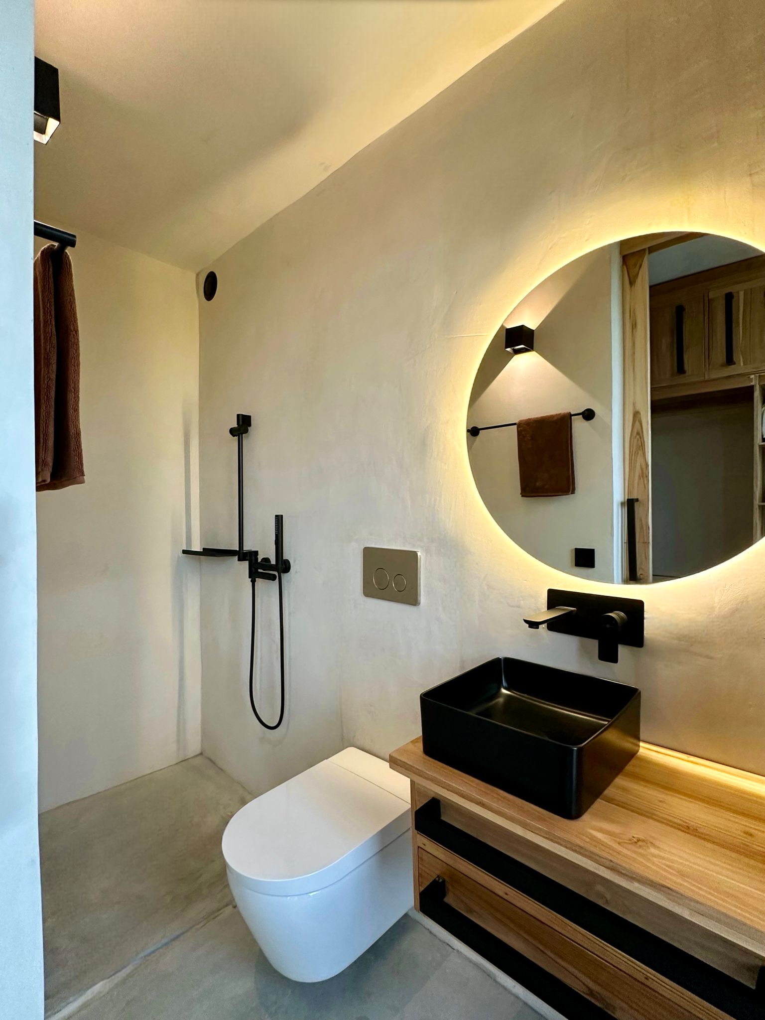 Finished bathroom with off-white microcement walls, grey microcement floor, round backlit mirror and black fixtures